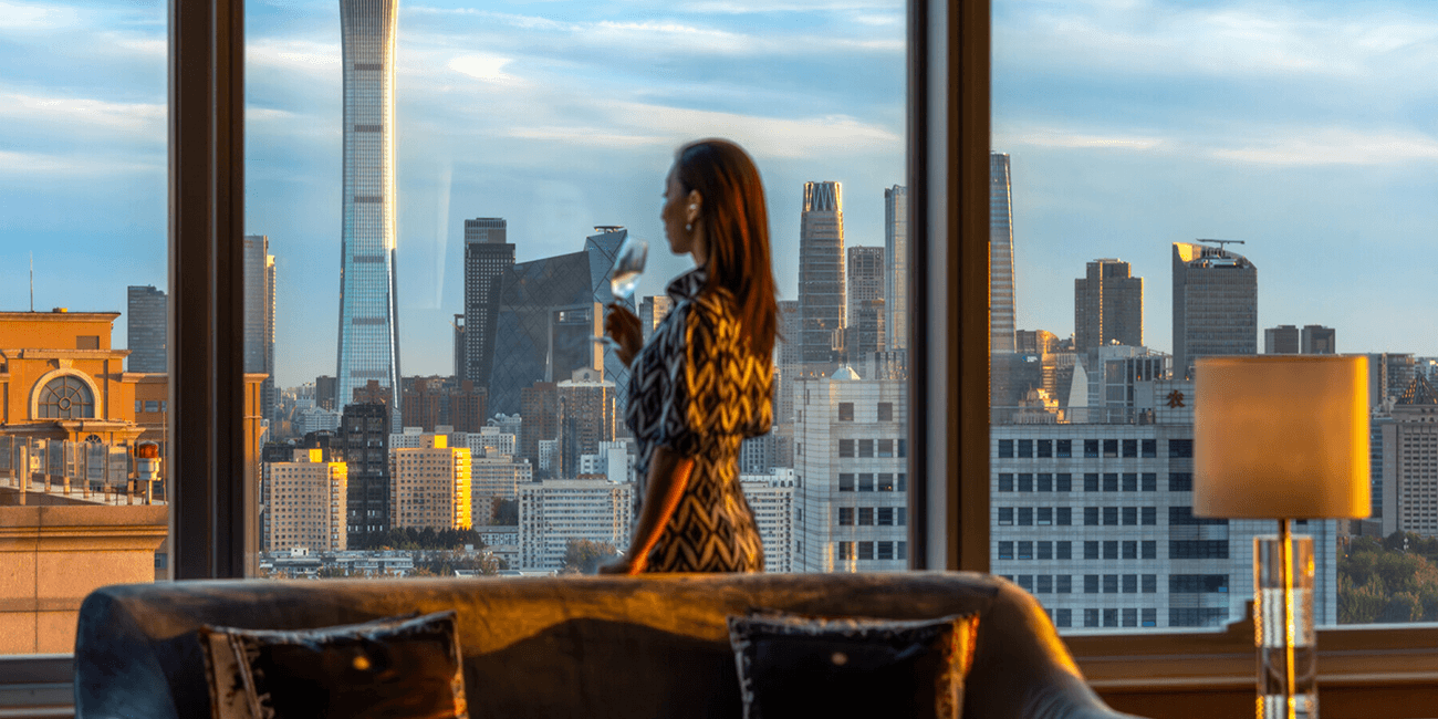 Woman holding a drink, gazing at a city skyline through a window.