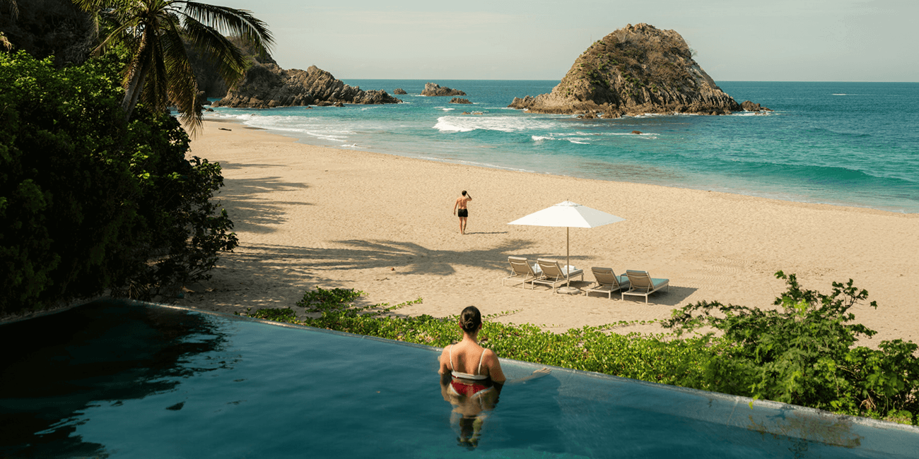Person relaxing in an infinity pool overlooking a beach and ocean.