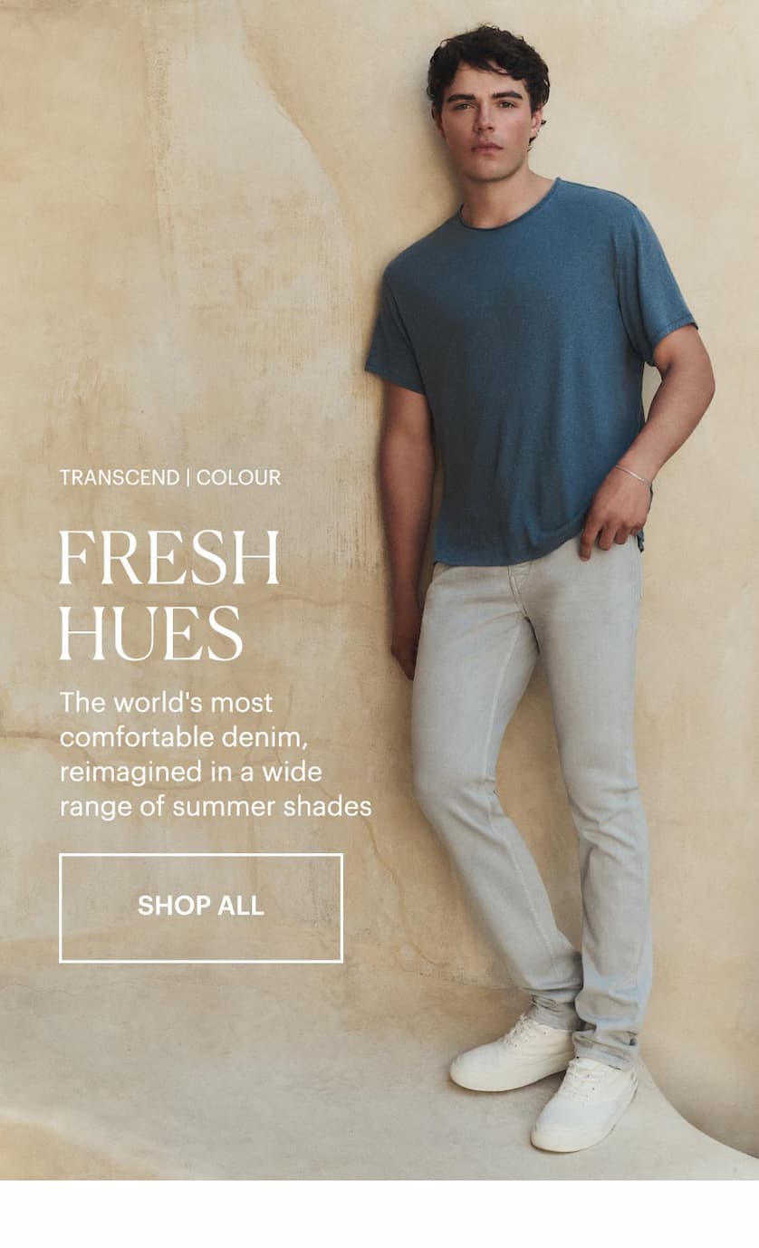 Transcend | Colour: Fresh Hues - The world's most comfortable denim, reimagined in a wide range of summer shades. Shop all. Man wearing a blue tee and light grey straight-leg jeans with white sneakers, standing against a beige sunlit stone wall.