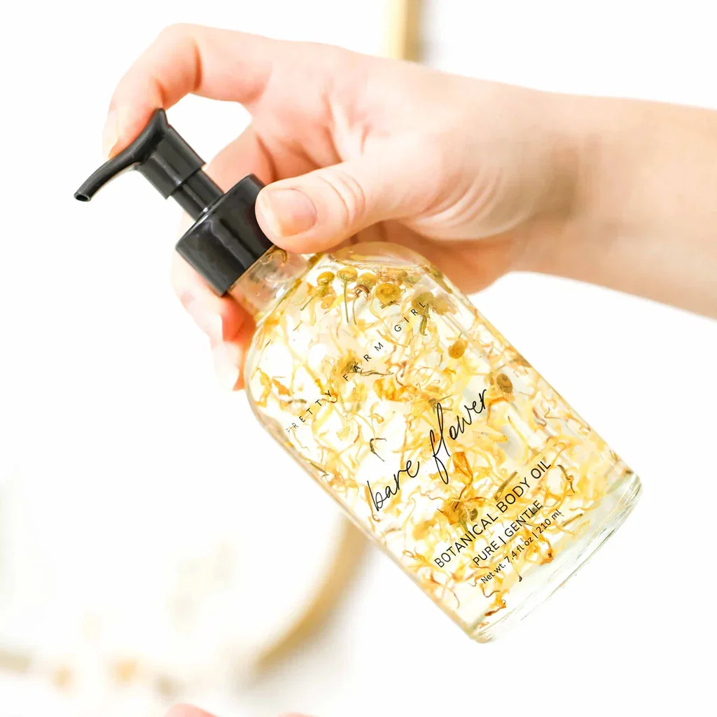 Person holding a bottle of Bare Flowers botanical body oil with gold leaf flakes and black pump dispenser.