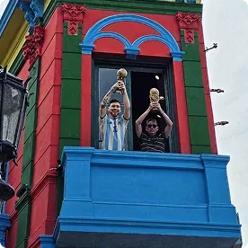 Two people standing on a bright blue balcony of a colorful red and green building, holding up trophies.
