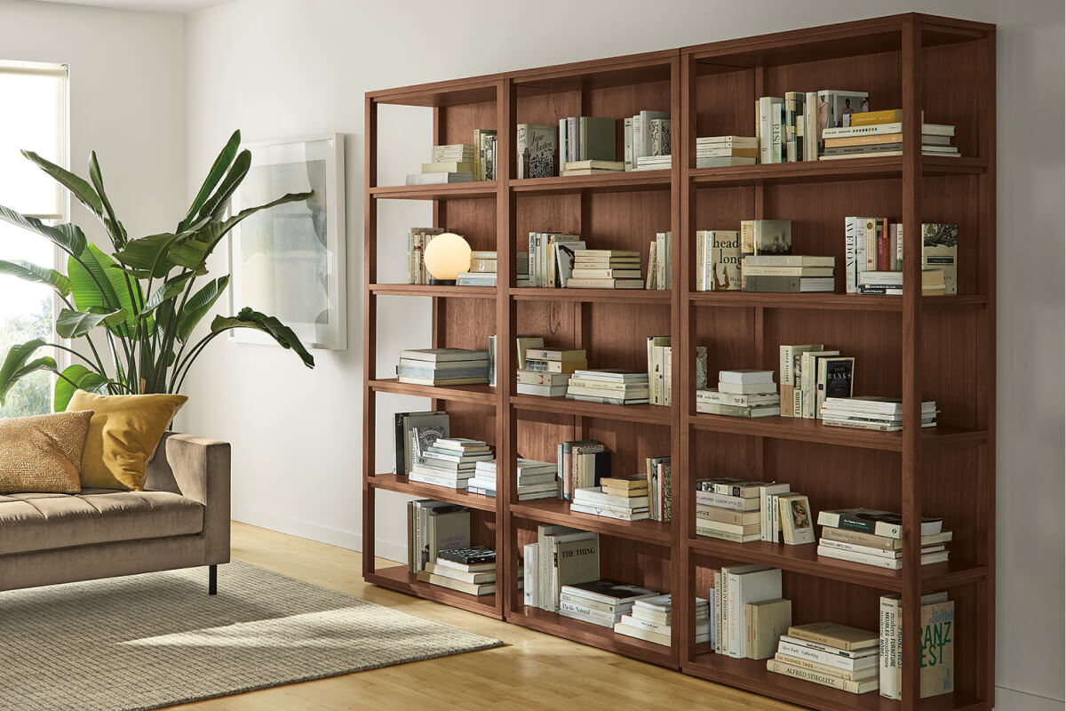 Fable walnut bookcases filled with stacked books beside a sofa, rug and large indoor plant.