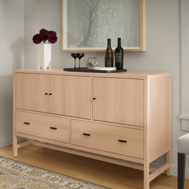 Berkeley cabinet in light wood with doors and drawers styled with wine bottles and framed artwork.