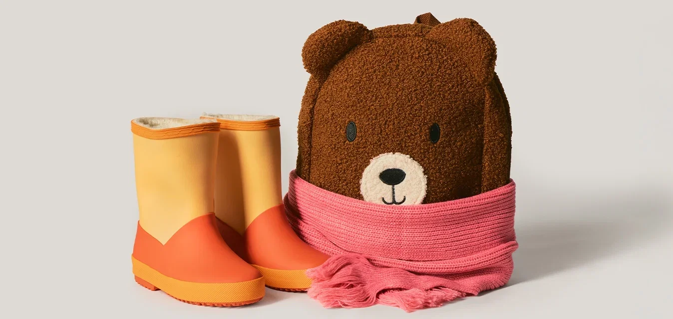 Kinderbruine pluche berenrugzak omwikkeld met een roze sjaal naast een paar oranje-gele regenlaarzen op een lichte achtergrond.