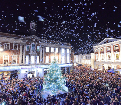 A large crowd gathers around a brightly lit Christmas tree, with snowflakes falling in a festive atmosphere.