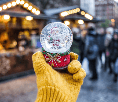 A person in a yellow glove holds a snow globe featuring Santa Claus, with festive market stalls in the background.