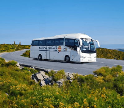A white coach with "National Holidays" branding driving along a scenic winding road surrounded by greenery.
