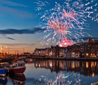 Fireworks illuminate the evening sky above a calm harbour, reflecting on the water.
