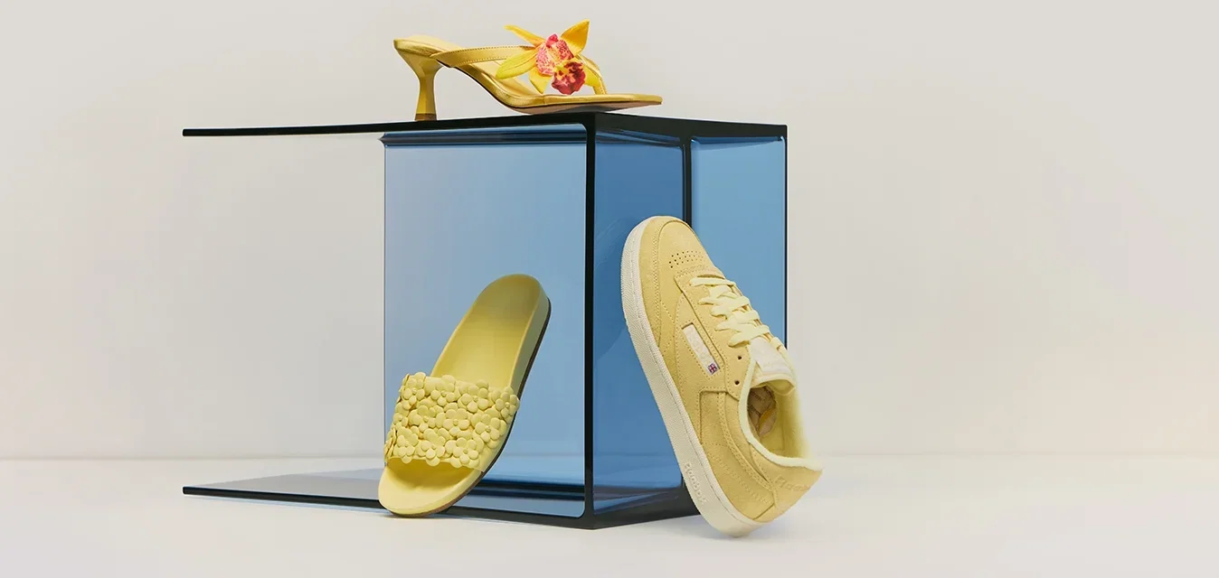 Trois chaussures jaunes — un talon aigu à motif floral, une mule texturée et une basket décontractée — présentées sur et autour d'un cube en verre bleu.