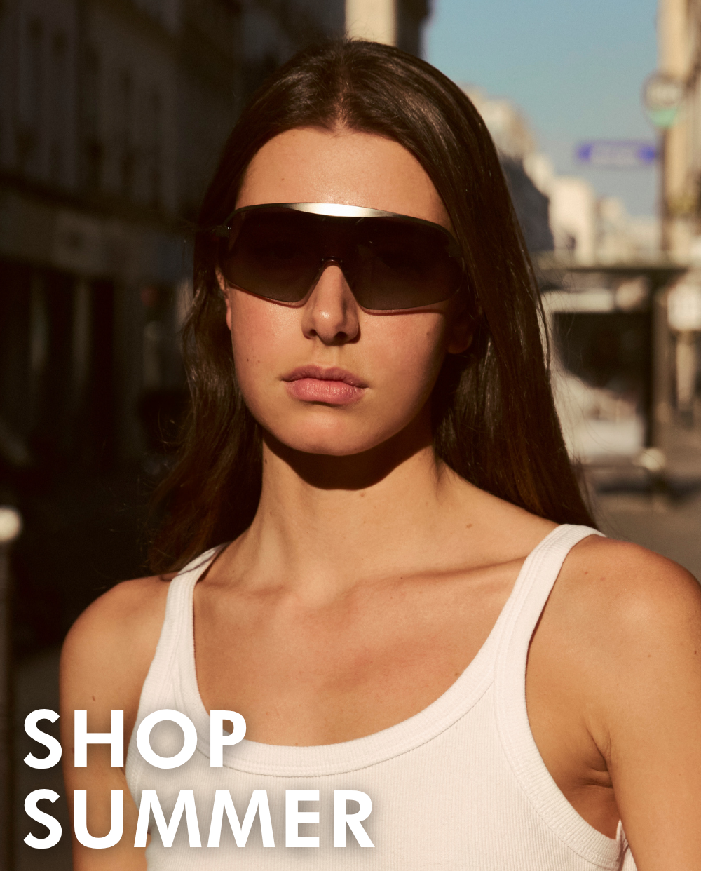 A portrait of a woman looking forward on a sunny city street, wearing a white tank top and the Hopper shield sunglasses with a silver top bar and gradient lenses. Bold white text in the lower-left corner reads, 