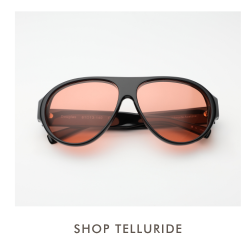 A studio product shot of the Telluride sunglasses, featuring an oversized black acetate aviator frame with a thick brow bar and deep orange-tinted lenses. Simple text below reads, 