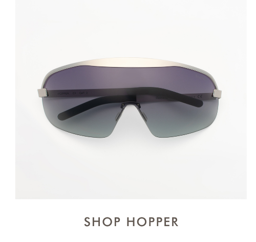 A studio product shot of the Hopper sunglasses, featuring a minimalist rimless shield design with a brushed silver top bar and a gradient lens transitioning from purple at the top to seafoam green at the bottom. Simple text below reads, 