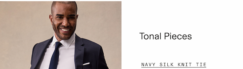 Man smiling in a navy suit, white dress shirt, and navy knit tie against a neutral background. "Tonal Pieces" "NAVY KNIT TIE"