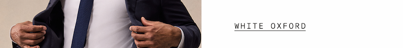  Close-up of a man's torso adjusting his navy blazer, wearing a navy knit tie and white dress shirt. "WHITE OXFORD"