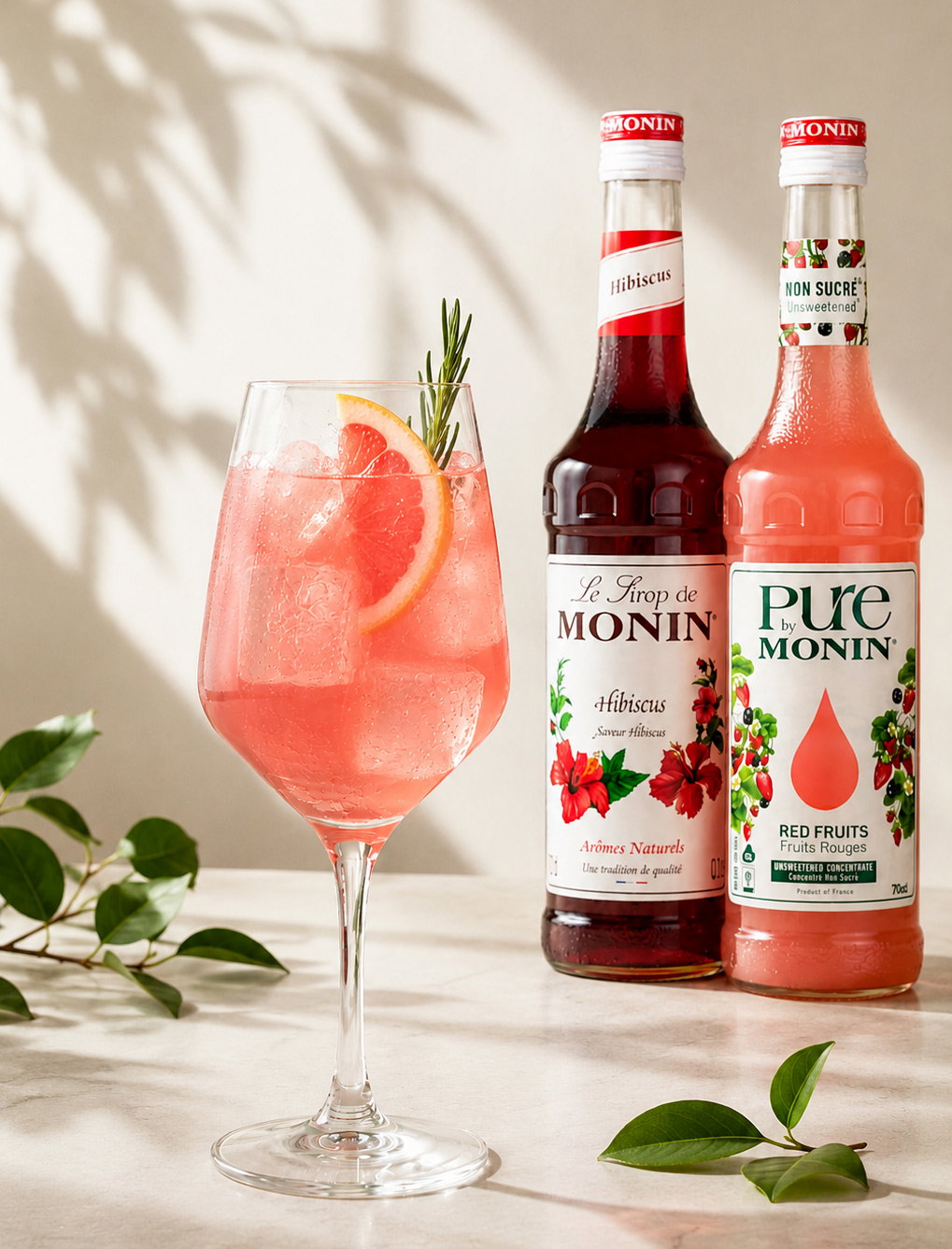Pink hibiscus drink with grapefruit slice and rosemary in a glass beside Monin Hibiscus and Red Fruits syrup bottles