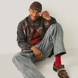 Junger Mann mit gemusterten Hosen, roten Socken, braunen Schuhen und einer Mütze, der lässig auf einem hellen Boden vor einer neutralen Wand sitzt.
