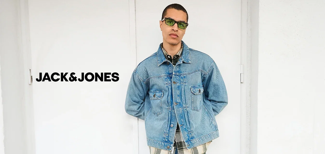 Mann mit grüner Sonnenbrille, hellblauer Jeansjacke über einem karierten Hemd, blauer Jeanshose und schwarzen Schuhen steht mit den Händen hinter dem Rücken an einer weißen Wand.