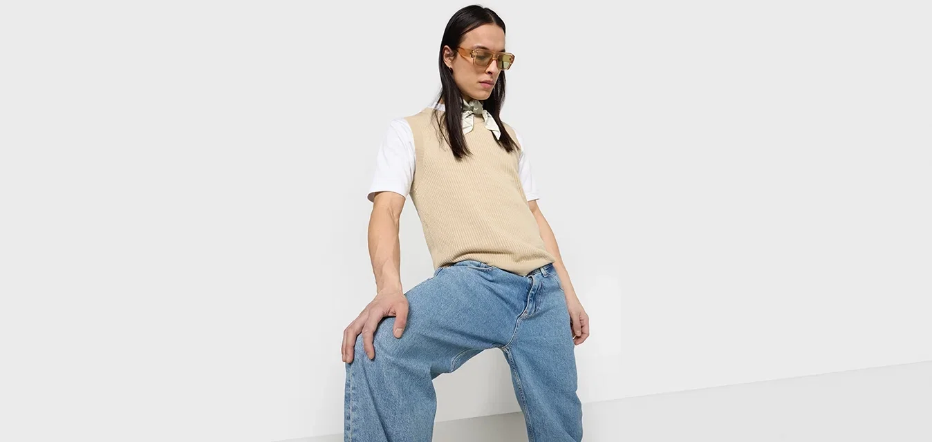 Beige gestrickte Weste über einem weißen T-Shirt, kombiniert mit hellblauen weiten Jeans. Die Accessoires umfassen gelbe Sonnenbrille und einen leichten Schal.