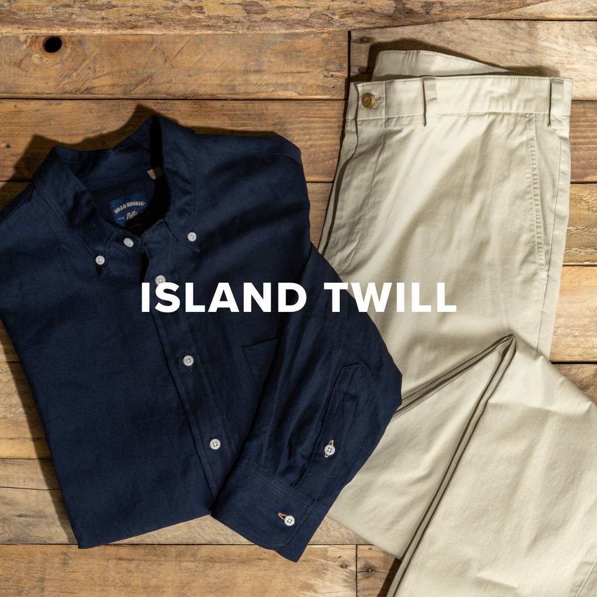 A pair of Bills Khakis Island Twill pants in Light Khaki color lay with a Bills Khakis Standard Fit Cotton Linen Shirt in Navy on a weathered wood surface