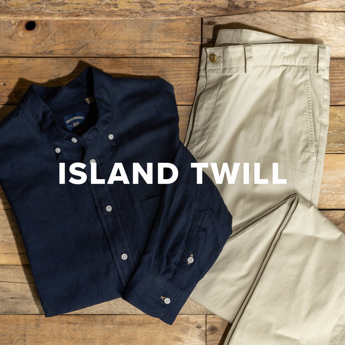 A pair of Bills Khakis Island Twill pants in Light Khaki color lay with a Bills Khakis Standard Fit Cotton Linen Shirt in Navy on a weathered wood surface