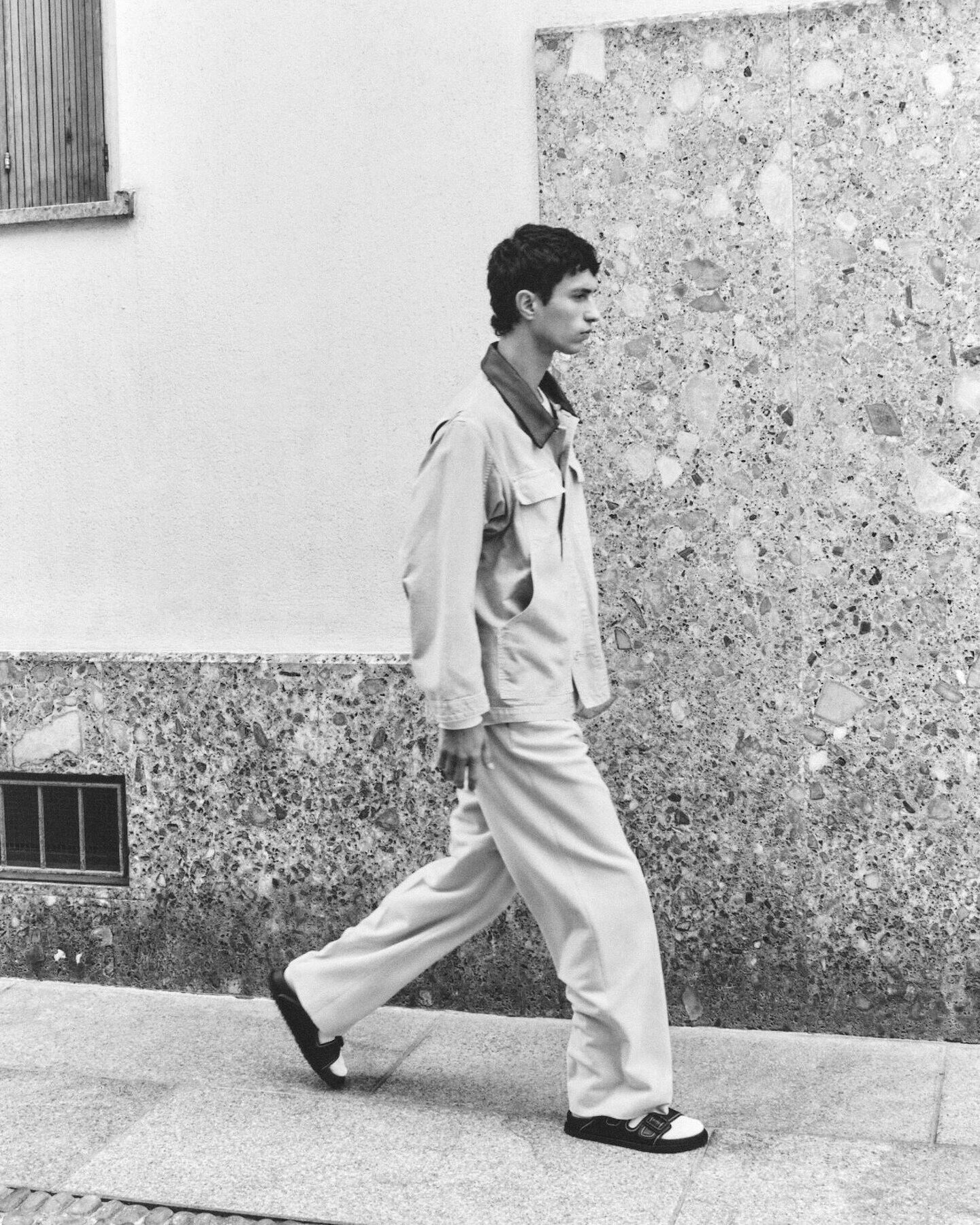 Black-and-white photo of a young man in a casual jacket and wide trousers walking past a textured stone wall in side profile