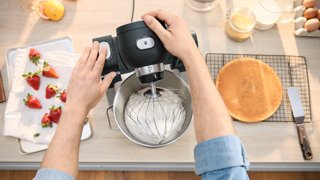 Mains fouettant un mélange avec un robot pâtissier Bosch. Fraises, gâteau et ingrédients de pâtisserie sur un comptoir.