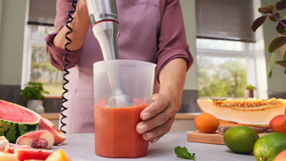 Mixeur plongeant Bosch utilisé pour un smoothie aux fruits (pastèque, pêches, melon, citrons verts) dans une cuisine lumineuse.