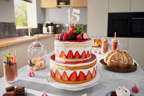 Gâteau d'anniversaire à deux étages (15 ans) avec fraises et baies, entouré de desserts variés sur un comptoir de cuisine.