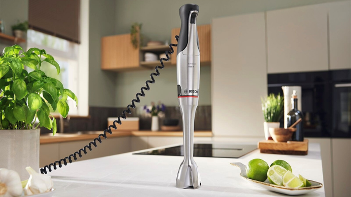 Mixeur plongeant Bosch en acier inoxydable sur un comptoir de cuisine moderne avec basilic, ail et citrons verts.
