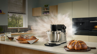 Robot pâtissier Bosch en action avec un nuage de farine, entouré de pains frais et d'ingrédients dans une cuisine moderne.