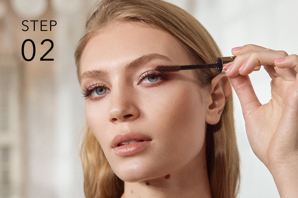 Beauty-Motiv mit „STEP 02“: Person trägt mit einer Mascara Bürste auf die Wimpern auf.