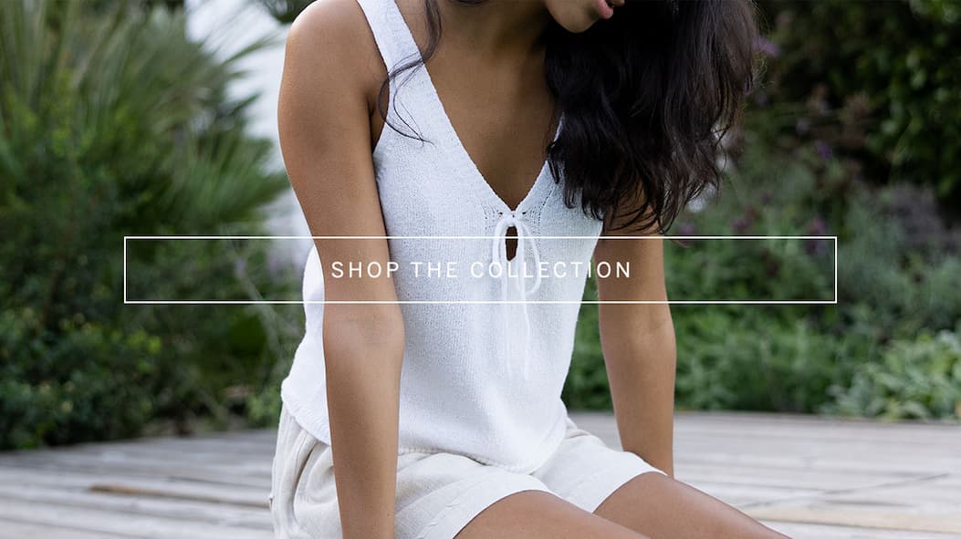 Close-up of a model sitting outdoors in a white sleeveless knit tank and light shorts, with overlay text reading “Shop the Collection.”