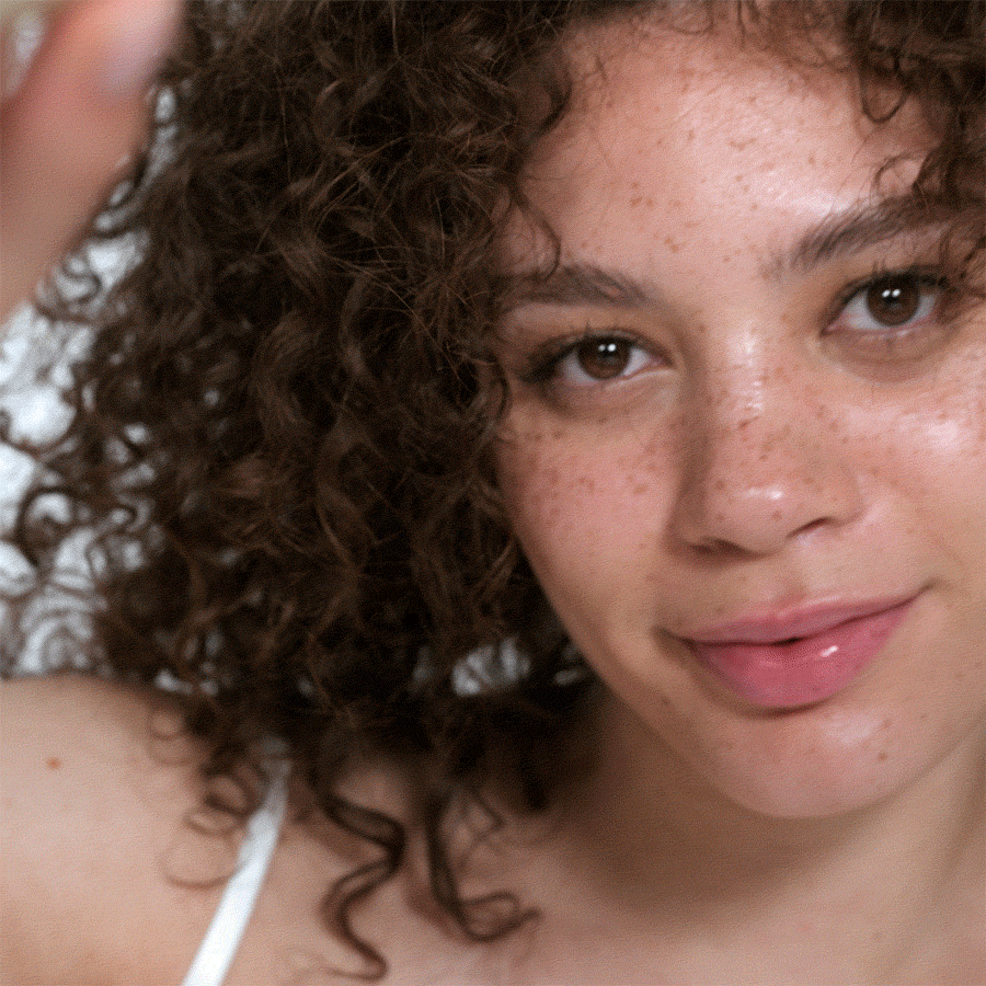A smiling woman with glowing, natural-looking skin holding a tube of bareMinerals Complexion Rescue tinted moisturizer close to her face.