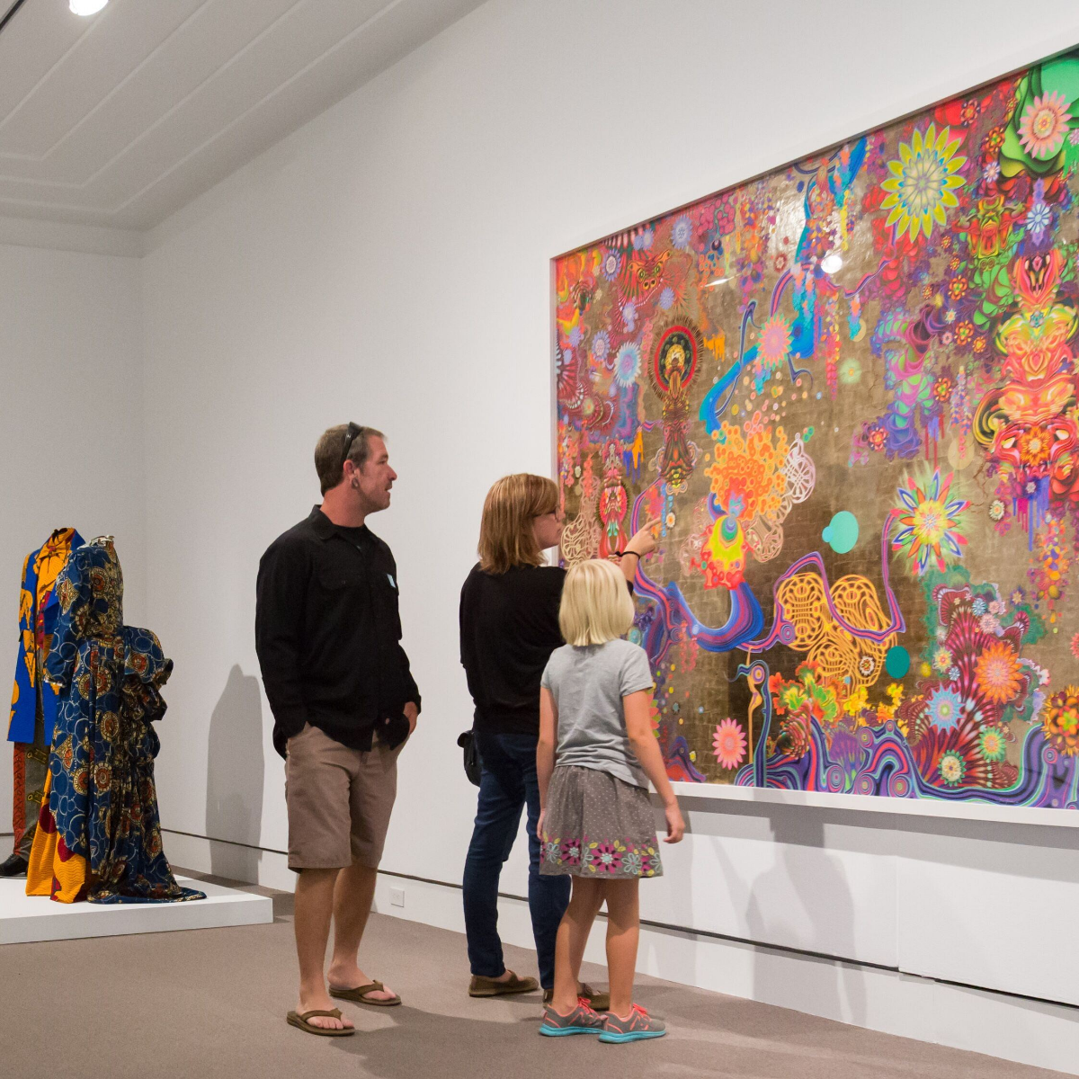A family stands together in an art gallery, closely observing a large, colorful abstract painting on the wall. The artwork features intricate patterns, swirling shapes, and bright, psychedelic colors. A sculptural textile piece with rich patterns and draped fabric is displayed on a pedestal nearby. The clean white walls and minimalist interior emphasize the vivid artwork and the viewers’ engagement.