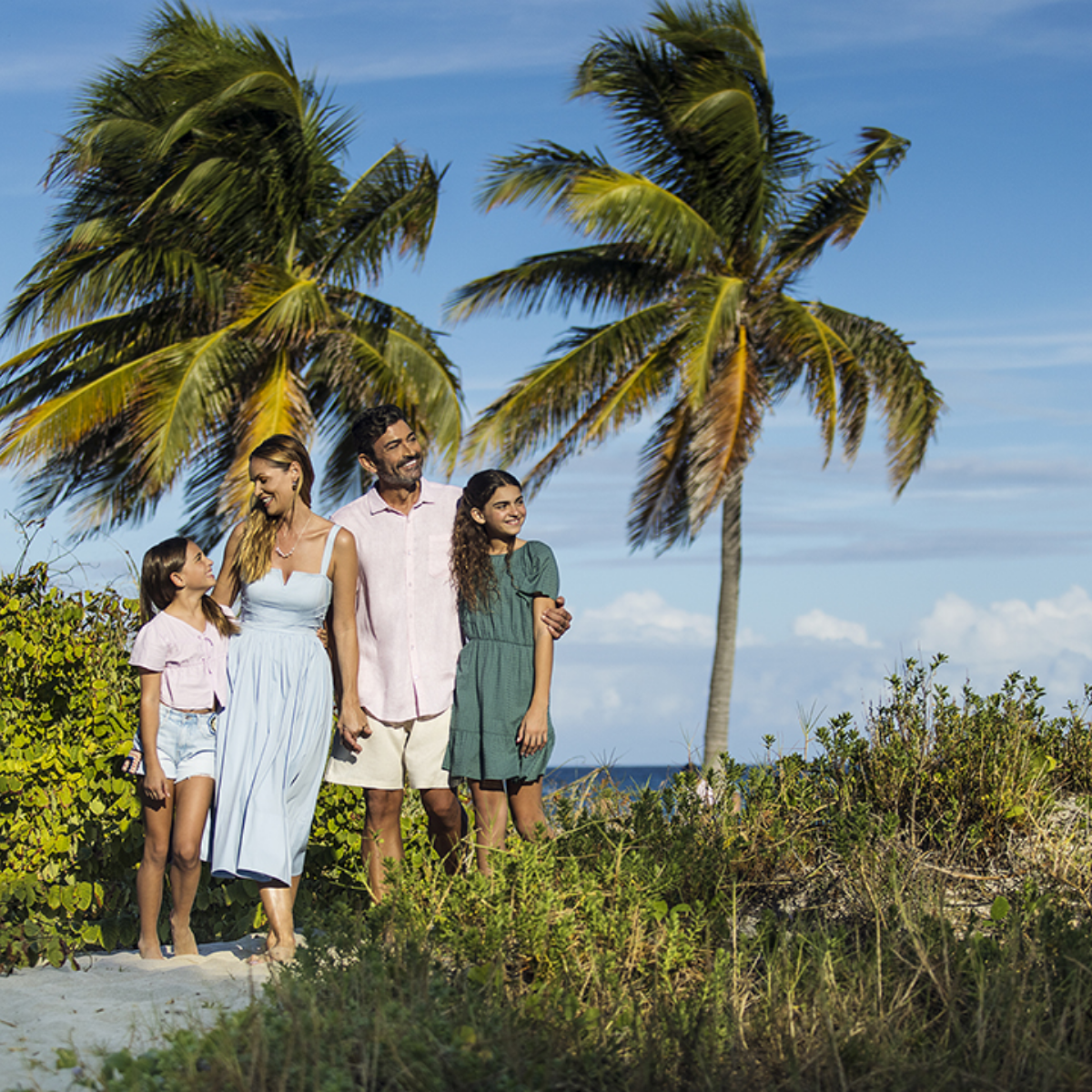 A family of four walks along a sandy path near the beach, surrounded by lush greenery and tall palm trees. The adults and children are dressed in light summer clothing, suggesting a warm, sunny day. The ocean and blue sky with scattered clouds are visible in the background, enhancing the tropical atmosphere. The scene conveys togetherness, relaxation, and a carefree vacation mood.