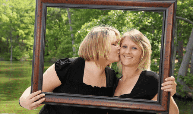 Two women are holding a large picture frame, with one kissing the other on the cheek. They are standing outdoors with greenery in the background.
