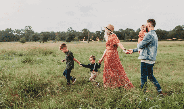 A family of five walks through a grassy field, holding hands. The father carries a small child, the mother wears a floral dress and hat, and two young boys walk beside them.