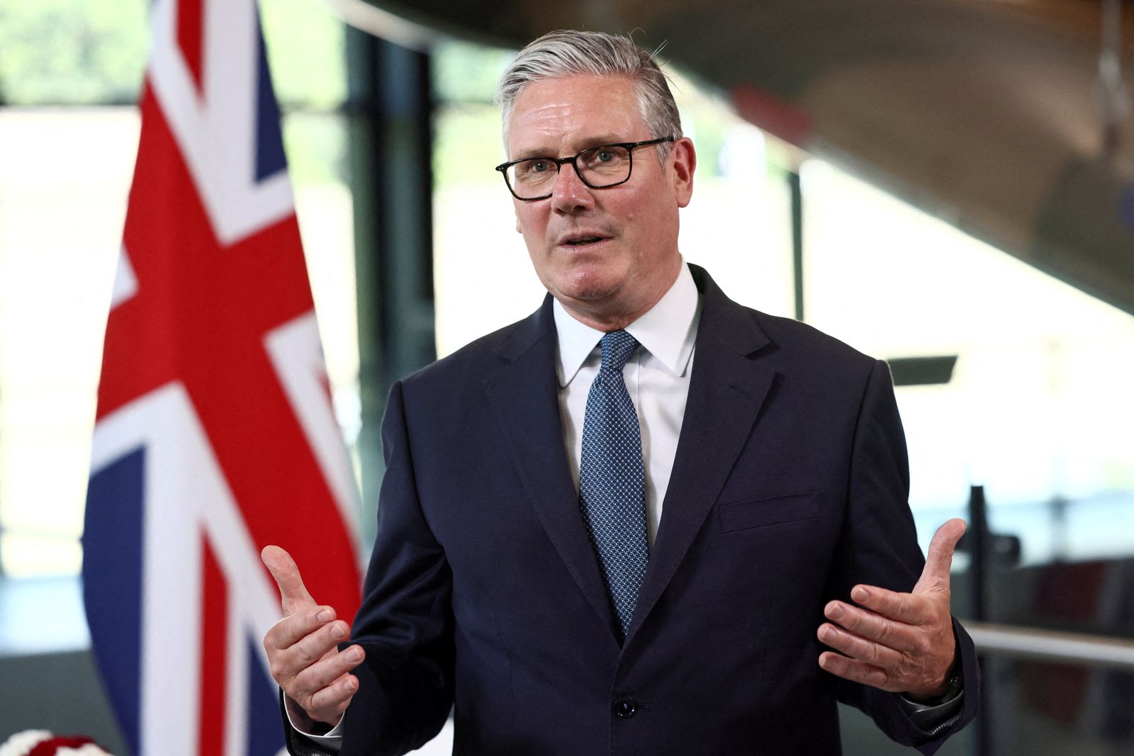 Keir Starmer stands in front of a Union flag blurred in the background. He raises his hands as he speaks. He is wearing a dark blue suit and a light blue tie.