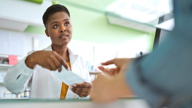 A woman wearing a long white pharmacist's coat hands someone wearing a blue shirt a prescription.