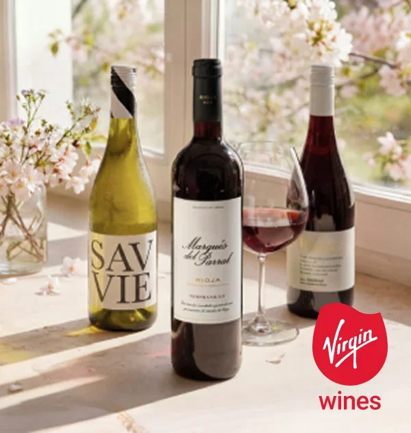 Three bottles of wine sit on a sunlit windowsill next to a filled red wine glass.