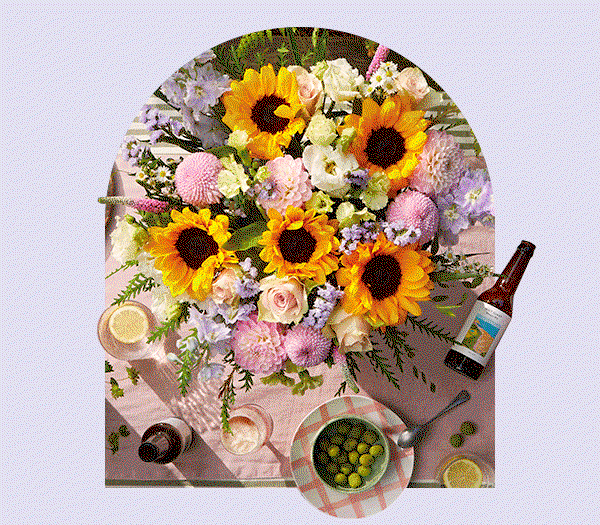 Bright summer bouquet featuring bold yellow sunflowers, soft pink dahlias, and delicate purple delphiniums, styled on a picnic table with drinks and olives.