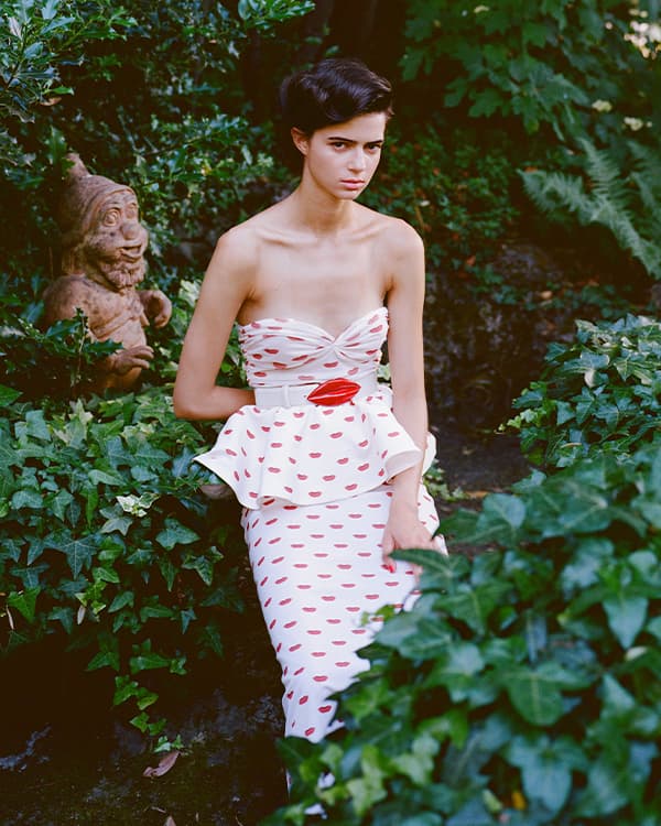Woman with short dark hair wearing a white strapless dress patterned with red lips, standing among lush green plants in a garden. 