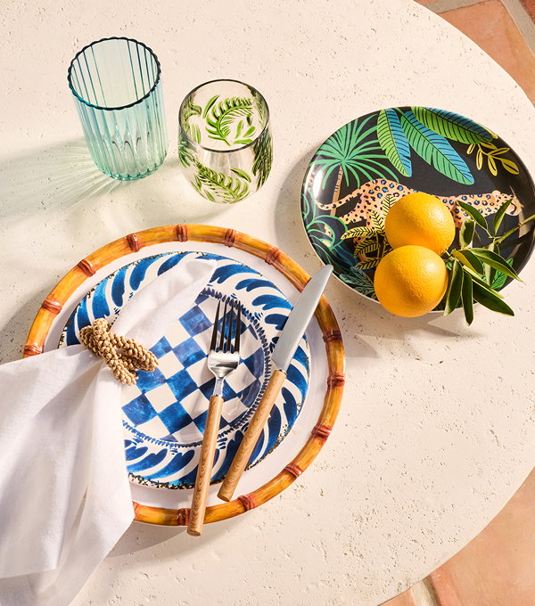 A tabletop place setting with patterned plates, wooden-handled cutlery, two glasses, and a bowl with lemons and greenery on a light surface.