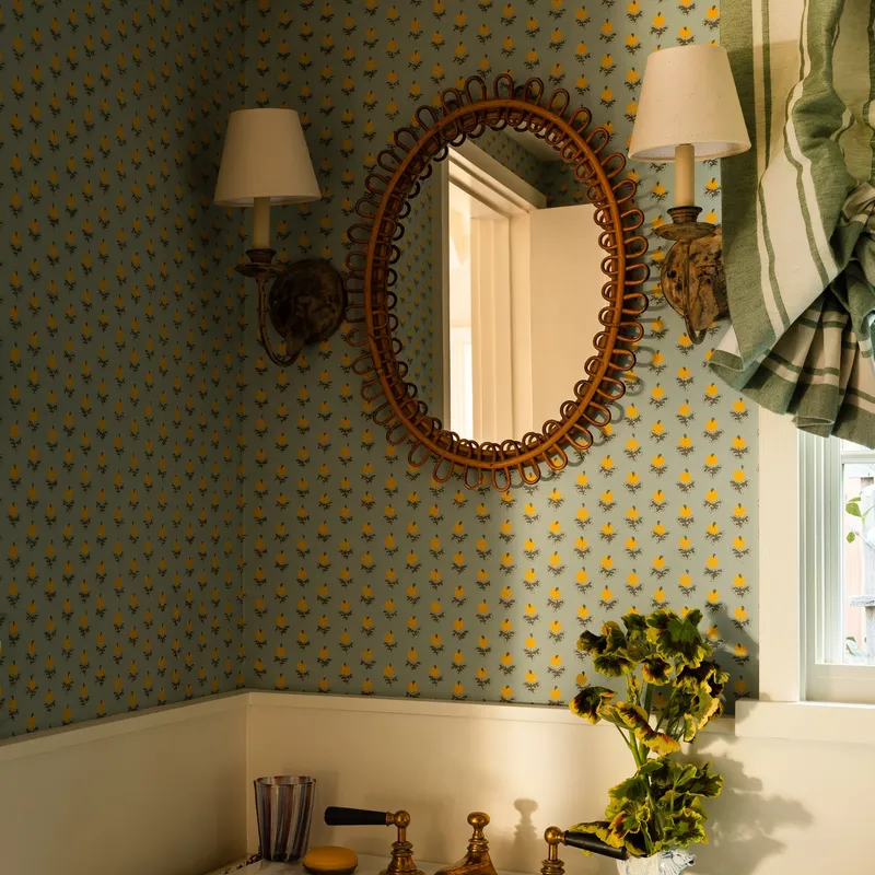  Wallpapered bathroom with an oval mirror that has a rattan frame surrounded by two sconces. 