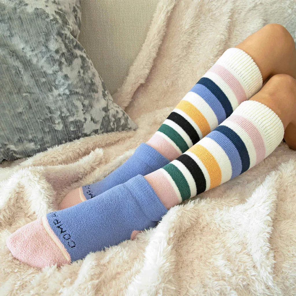Two pairs of colorful striped Comrad Socks displayed on legs against a cozy cream blanket backdrop.
