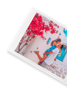 A polaroid-style photo of a couple embracing outside a whitewashed building with vibrant red bougainvillea, on a soft teal background.