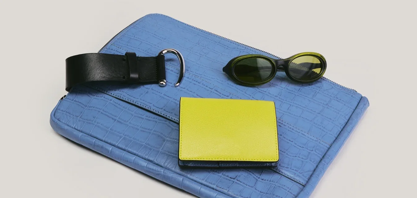 Blauer strukturierter Clutch mit schwarzem Lederband, gelbem Portemonnaie und dunkler ovaler Sonnenbrille auf hellem Hintergrund angeordnet.