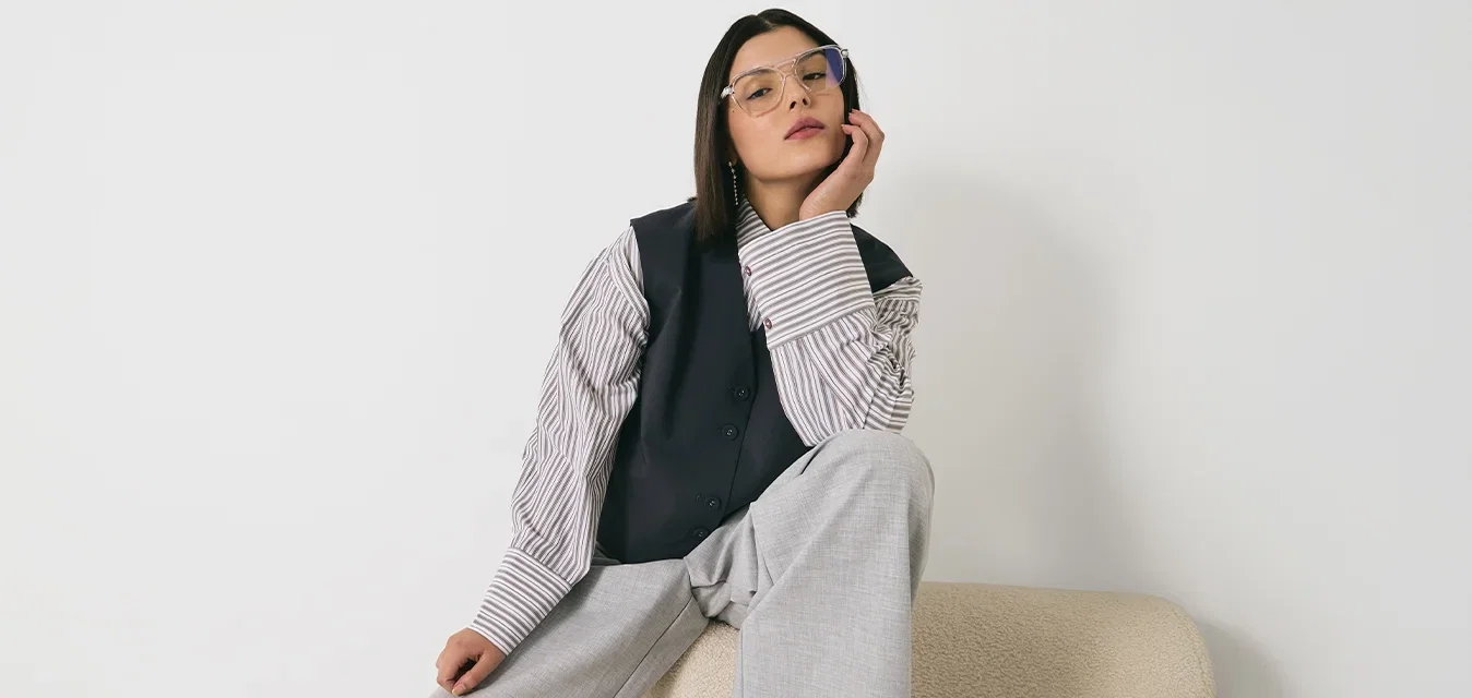 Übergroße graue Hose, schwarze Weste über einem gestreiften Hemd, Brille mit hellblauen Rahmen. Sitzt auf einem beigefarbenen strukturierten Sofa vor einer weißen Wand.