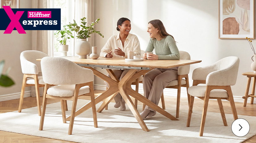 Ein großes, helles Ecksofa mit strukturierter Oberfläche steht in einem modern eingerichteten Wohnzimmer. Zwei Personen sitzen entspannt nebeneinander auf dem Sofa, eine Person hält ein Buch, die andere eine Tasse. Vor dem Sofa steht ein runder, dreistufiger Glastisch mit Metallgestell, darauf eine Vase mit Zweigen. Im Hintergrund sind große Fenster mit hellen Vorhängen und ein abstraktes Wandbild zu sehen. Links oben befindet sich ein auffälliges, mehrfarbiges Logo mit dem Schriftzug 'Höffner express'.