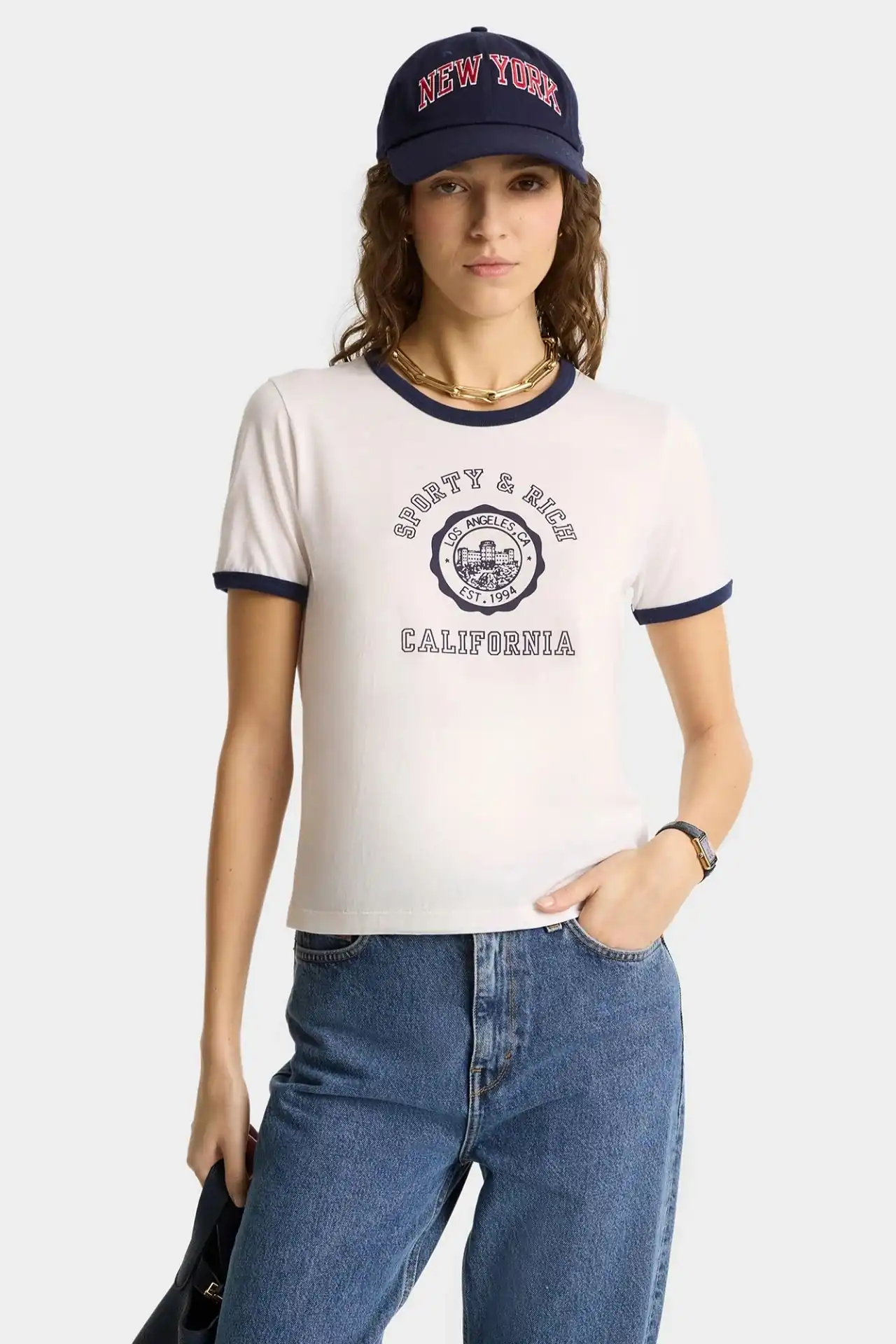 A medium shot shows a woman wearing a white ringer t-shirt with navy trim, blue jeans, and a navy baseball cap that reads 'NEW YORK' in red lettering,...